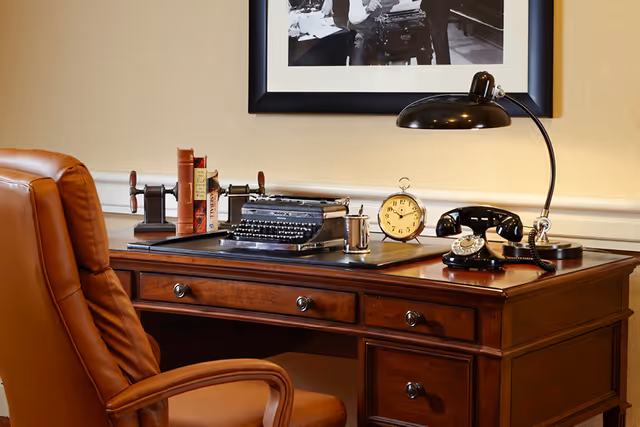 A classic wooden desk with a brown leather office chair. On the desk are vintage items including a typewriter, an old-fashioned black rotary telephone, a round alarm clock, a desk lamp, a metal cup, and a few books held by a bookend. A black and white framed photograph hangs on the beige wall behind the desk.