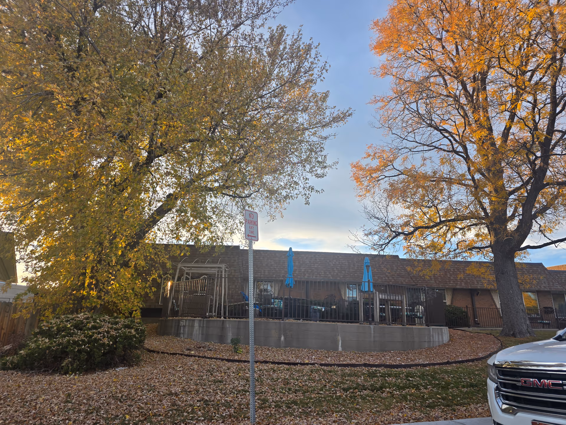 Outdoor view of Clear Creek Care Center showing a patio area with blue umbrellas, surrounded by a metal fence. The scene is set in autumn with trees having yellow and orange leaves and fallen leaves covering the ground. A 'No Parking Any Time' sign is visible in front of the patio, and part of a white GMC vehicle is seen on the right side.