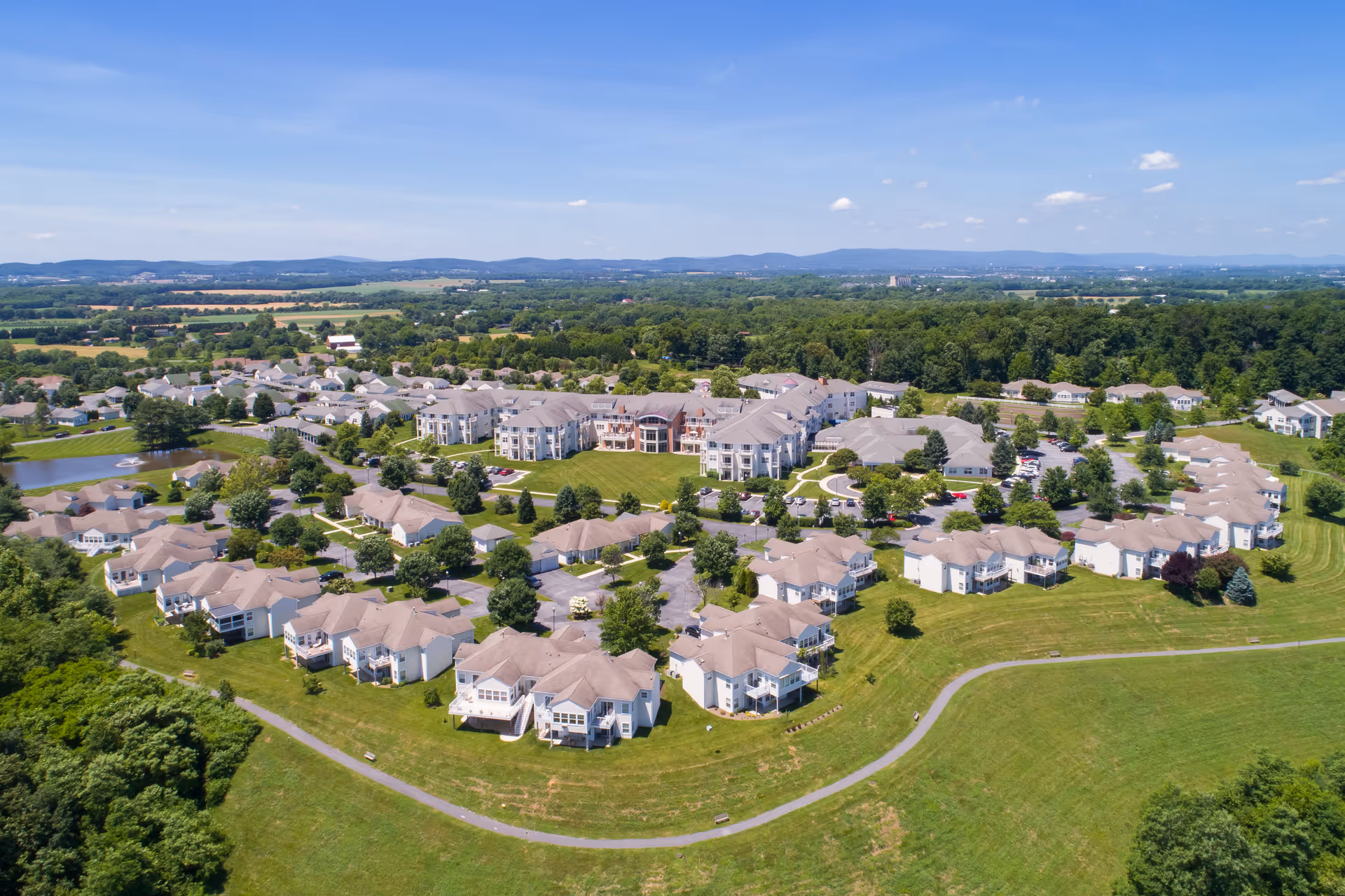 Aerial view of Buckingham's Choice senior living community showing multiple residential buildings with beige roofs surrounded by green lawns, trees, and a walking path under a clear blue sky.