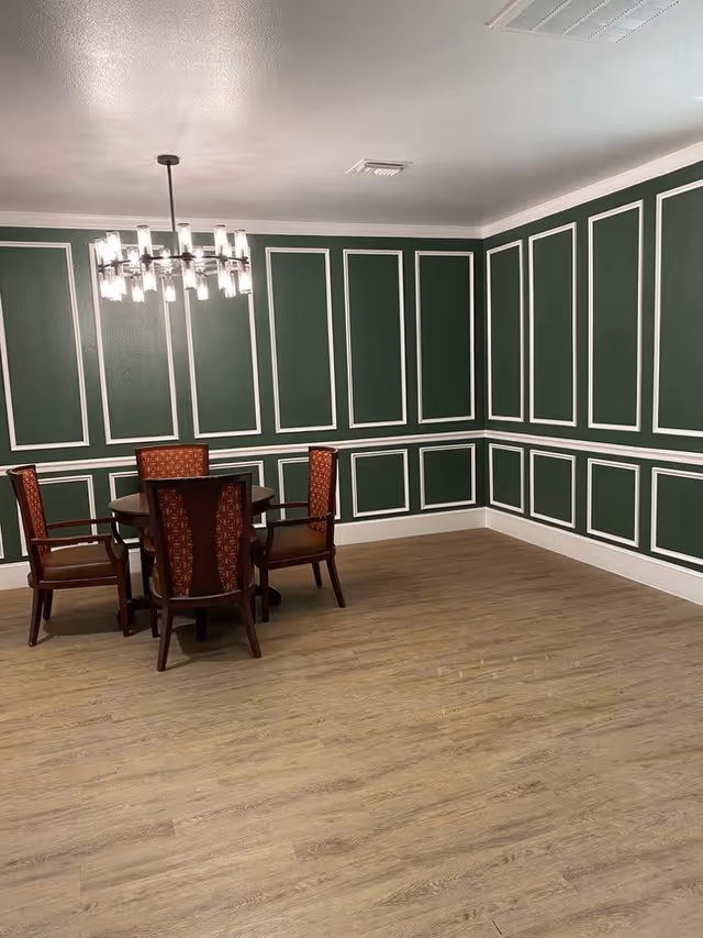 A dining area with a round wooden table and four wooden chairs with red patterned upholstery. The walls are painted dark green with white decorative molding, and the floor is light wood. A modern chandelier with multiple lights hangs above the table.