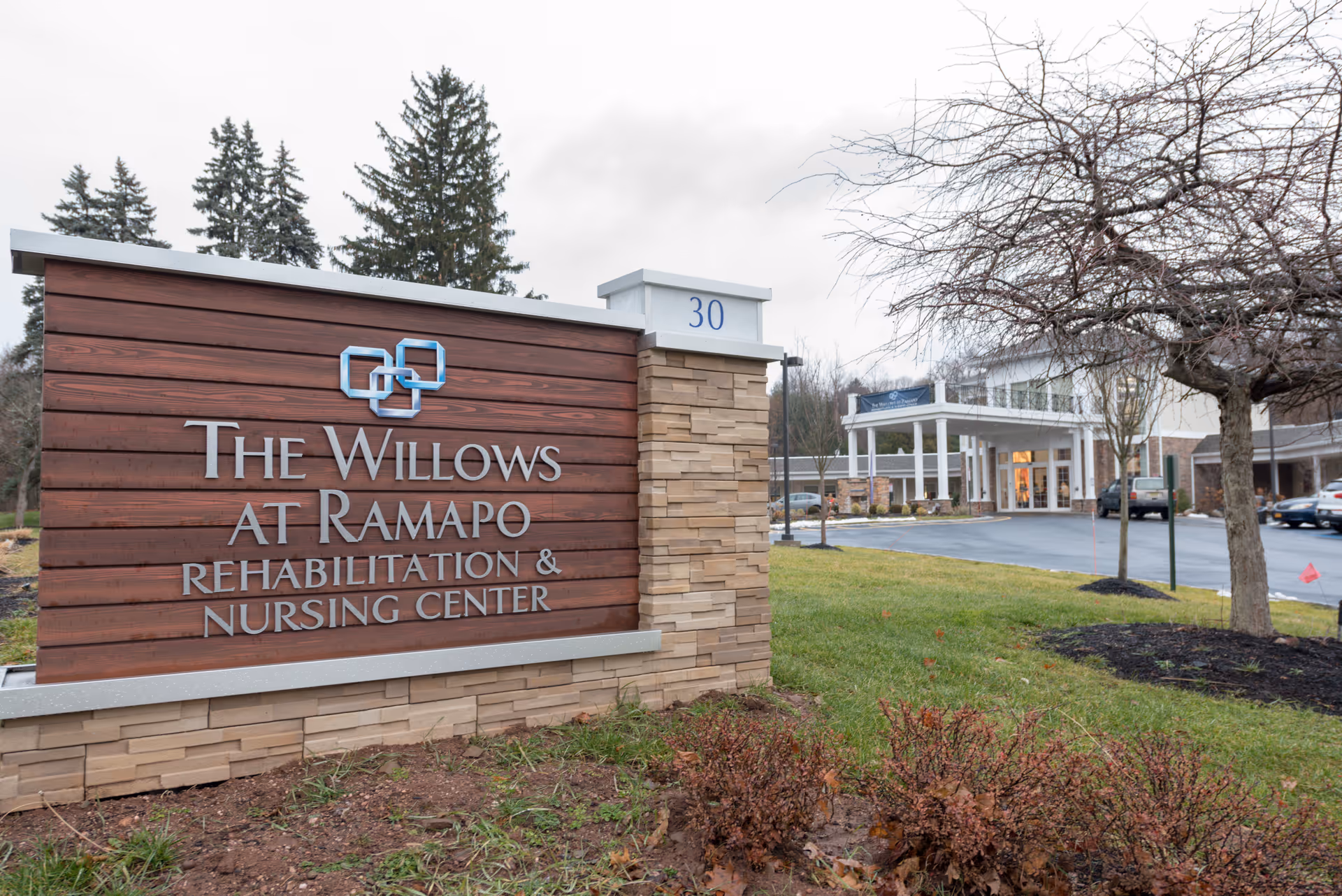 Outdoor view of the entrance to The Willows at Ramapo Rehabilitation & Nursing Center, featuring a large wooden and stone sign with the facility name and the number 30. The building entrance and parking area are visible in the background with some trees and landscaping around.