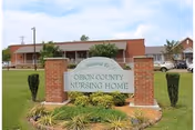 Exterior view of Obion County Nursing Home building with a brick sign in the foreground that reads 'Welcome to Obion County Nursing Home'. The building is a single-story structure with a red roof and several windows. There is a well-maintained lawn and some shrubs around the sign.