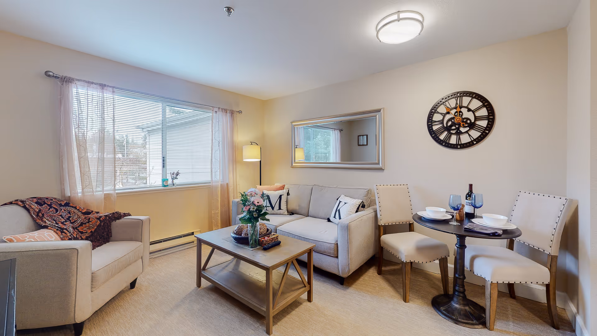 A cozy living room area with a beige armchair and a beige sofa adorned with decorative pillows. A wooden coffee table with a vase of flowers and decorative items sits in front of the sofa. To the right, there is a small round dining table set for two with white bowls, blue glasses, and a bottle of wine. A large window with sheer curtains lets in natural light, and a wall clock and a rectangular mirror hang on the walls.