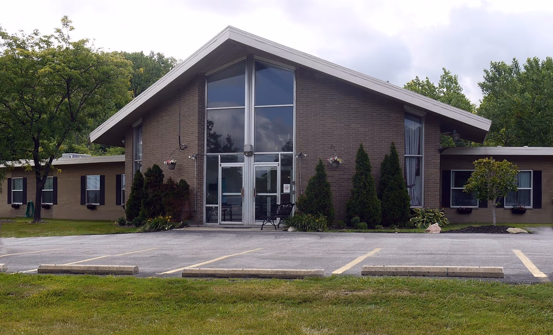 Front exterior view of a single-story brick building with a large triangular roof and glass entrance doors. The building is surrounded by trees and shrubs, with a parking lot in front featuring several empty parking spaces.