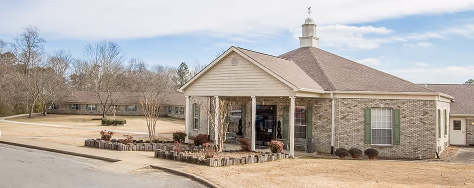 Exterior view of a single-story brick building with a peaked roof and a small cupola on top. The entrance has a covered porch supported by white columns, with some landscaping including small bushes and trees around the building. The sky is partly cloudy.