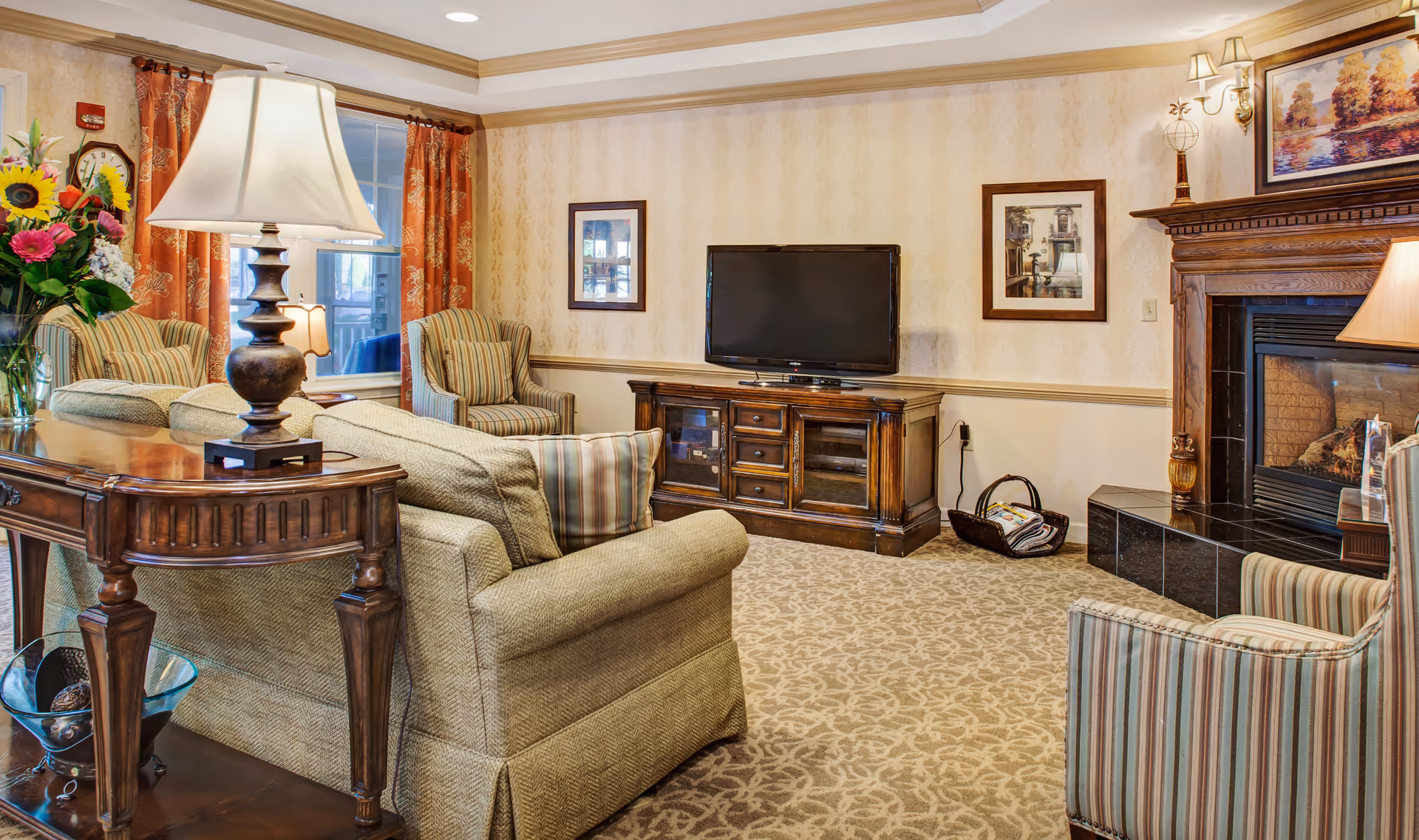 Bright, traditionally furnished communal living room with sofas and armchairs, a TV on a wooden console, and a fireplace.