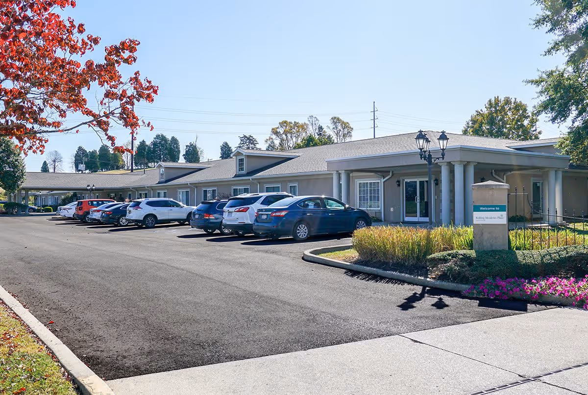 Exterior view of Rolling Meadows Place facility with a parking lot filled with cars, a building with a covered entrance supported by columns, and landscaped areas with grass, flowers, and trees under a clear blue sky.
