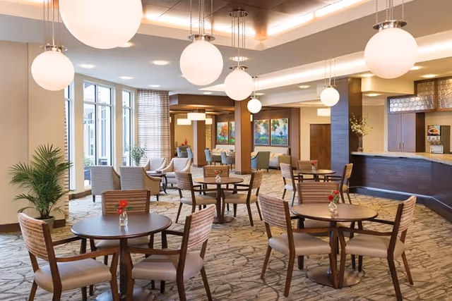 A spacious and well-lit common area with round wooden tables and chairs arranged for seating. Large spherical pendant lights hang from the ceiling. There are large windows on one side letting in natural light, and a counter area with cabinets on the right. The room has a carpeted floor with a patterned design, some potted plants, and colorful artwork on the walls in the background.