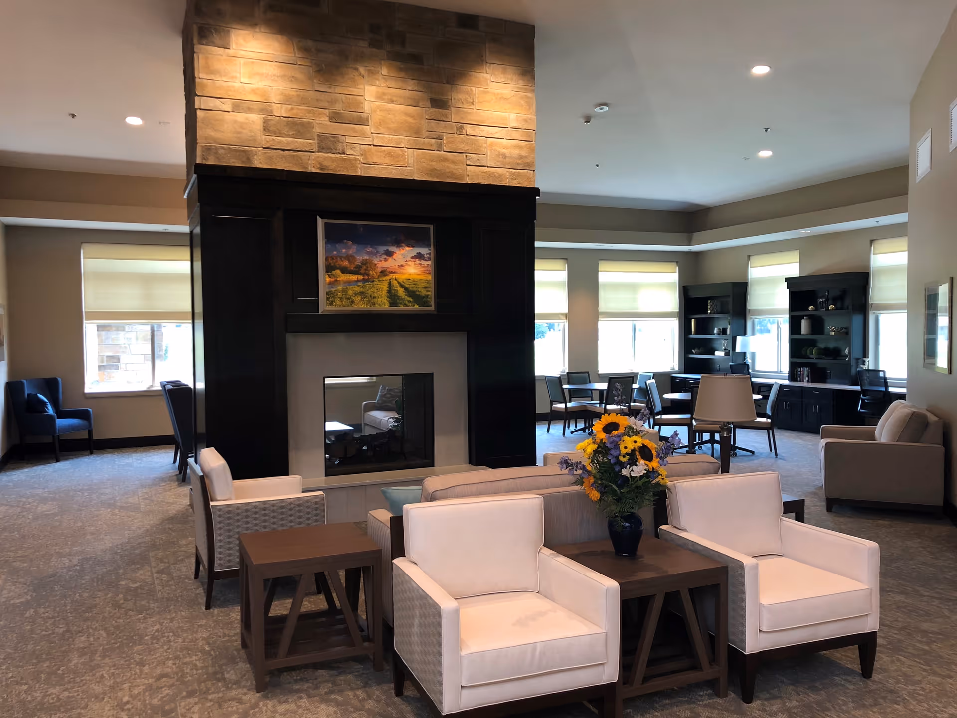 A spacious senior living facility common area with a central stone fireplace. The room features comfortable seating including white armchairs and sofas arranged around wooden tables. Large windows with blinds allow natural light to fill the room. There are bookshelves and additional seating areas in the background, with a vase of colorful flowers on one of the tables.