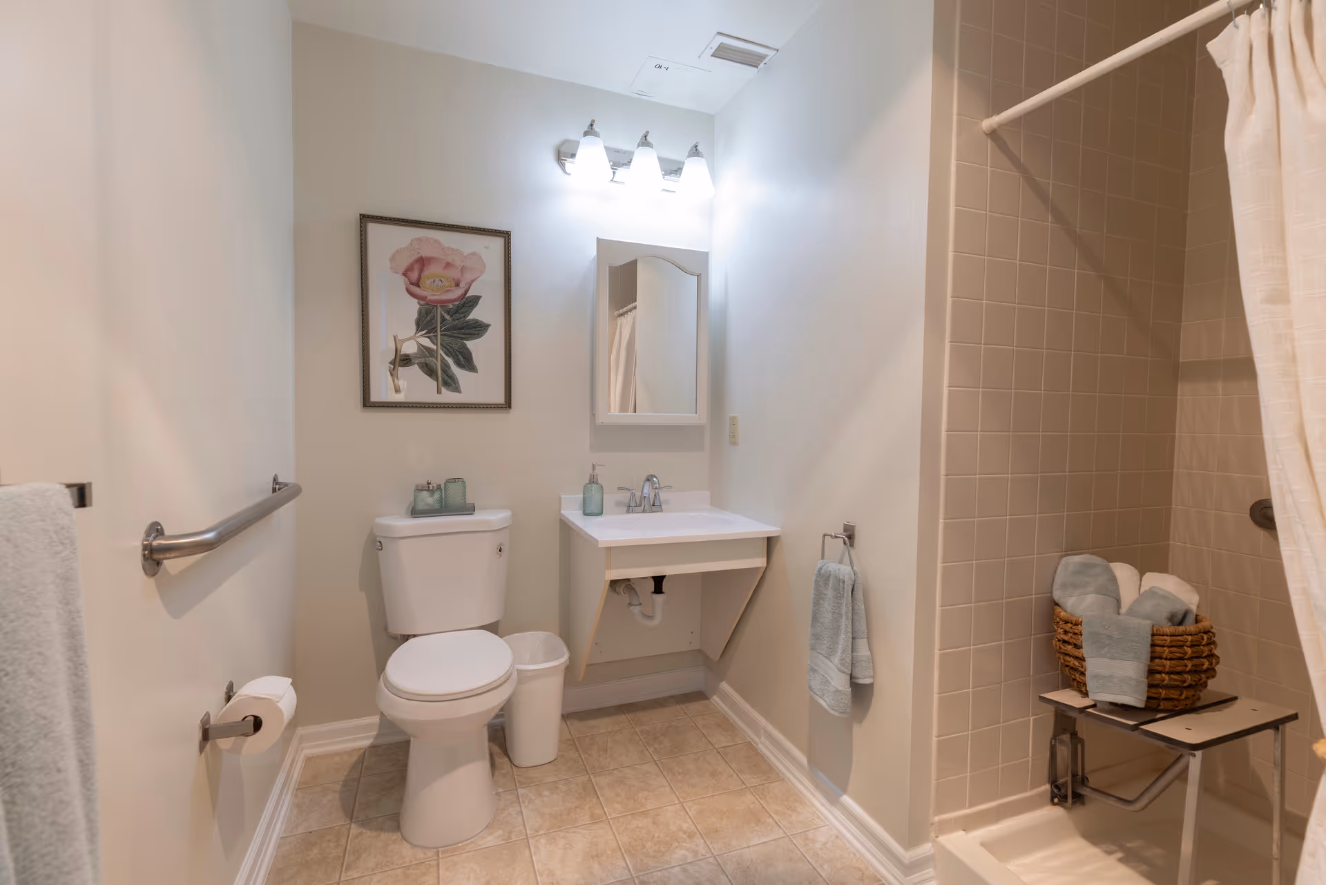 A clean and well-lit bathroom featuring a toilet with a small trash bin beside it, a wall-mounted sink with a mirror and light fixture above, a framed floral artwork on the wall, and a tiled shower area with a shower curtain and a basket holding rolled towels on a fold-down shower seat.