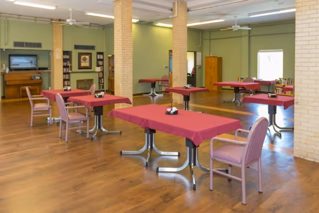 A spacious dining room with wooden floors and light green walls. Several tables covered with red tablecloths are arranged throughout the room, each accompanied by pink cushioned chairs. The room features brick pillars, ceiling fans, and a piano against one wall with framed pictures and shelves nearby.