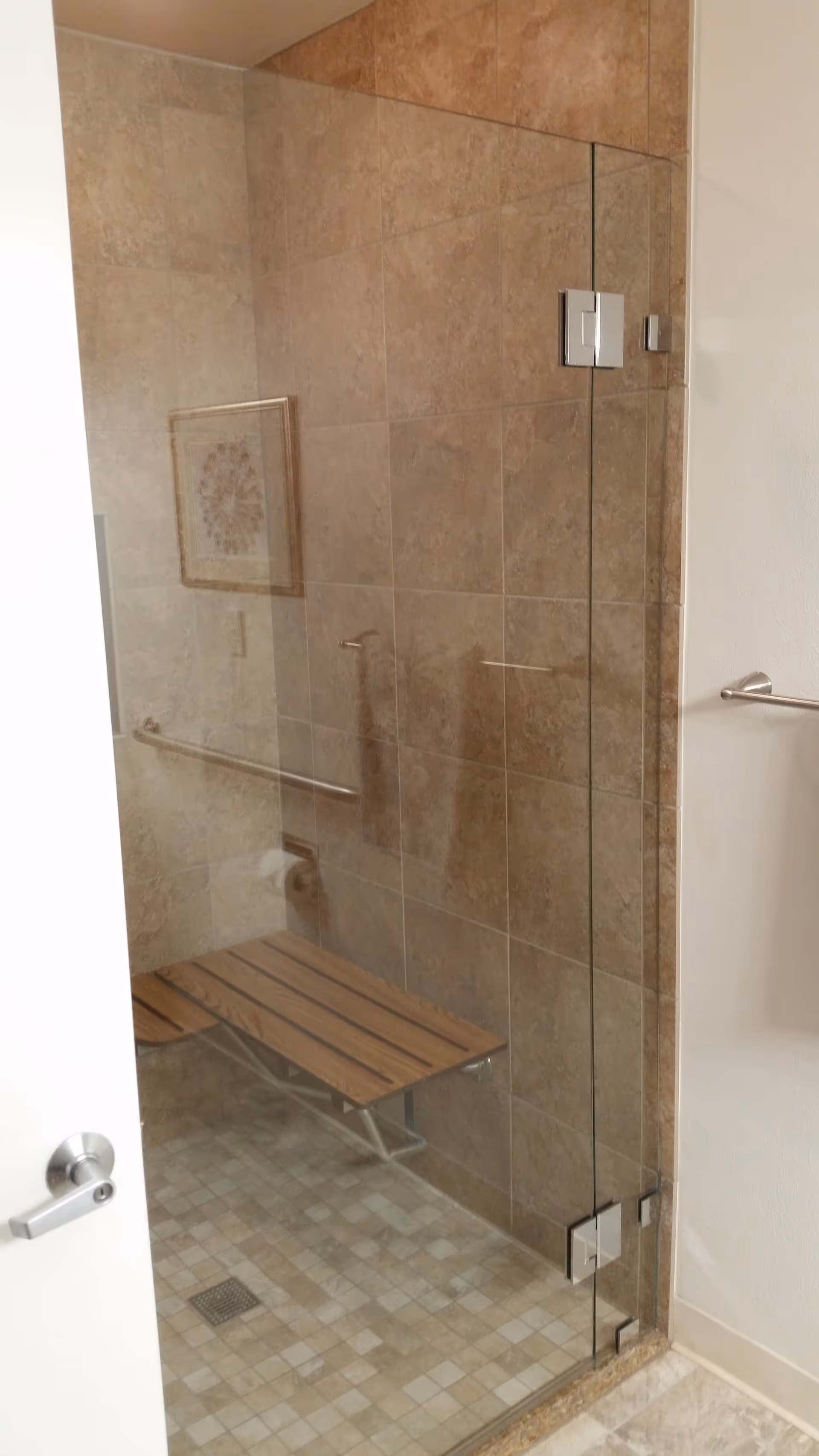 A tiled shower area with a glass door, featuring a wooden bench attached to the wall and a metal grab bar. The walls and floor are covered with beige tiles, and there is a towel bar mounted on the adjacent wall outside the shower.