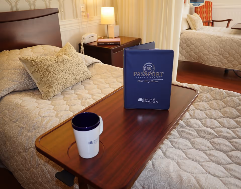 A neatly made bed with a beige patterned quilt and decorative pillow in a senior living facility room. A wooden overbed table holds a white mug and a blue folder labeled 'Passport Rehabilitation Your Way Home' from National Health Care. In the background, there is a nightstand with a lamp, a telephone, and a book, as well as another bed partially visible.