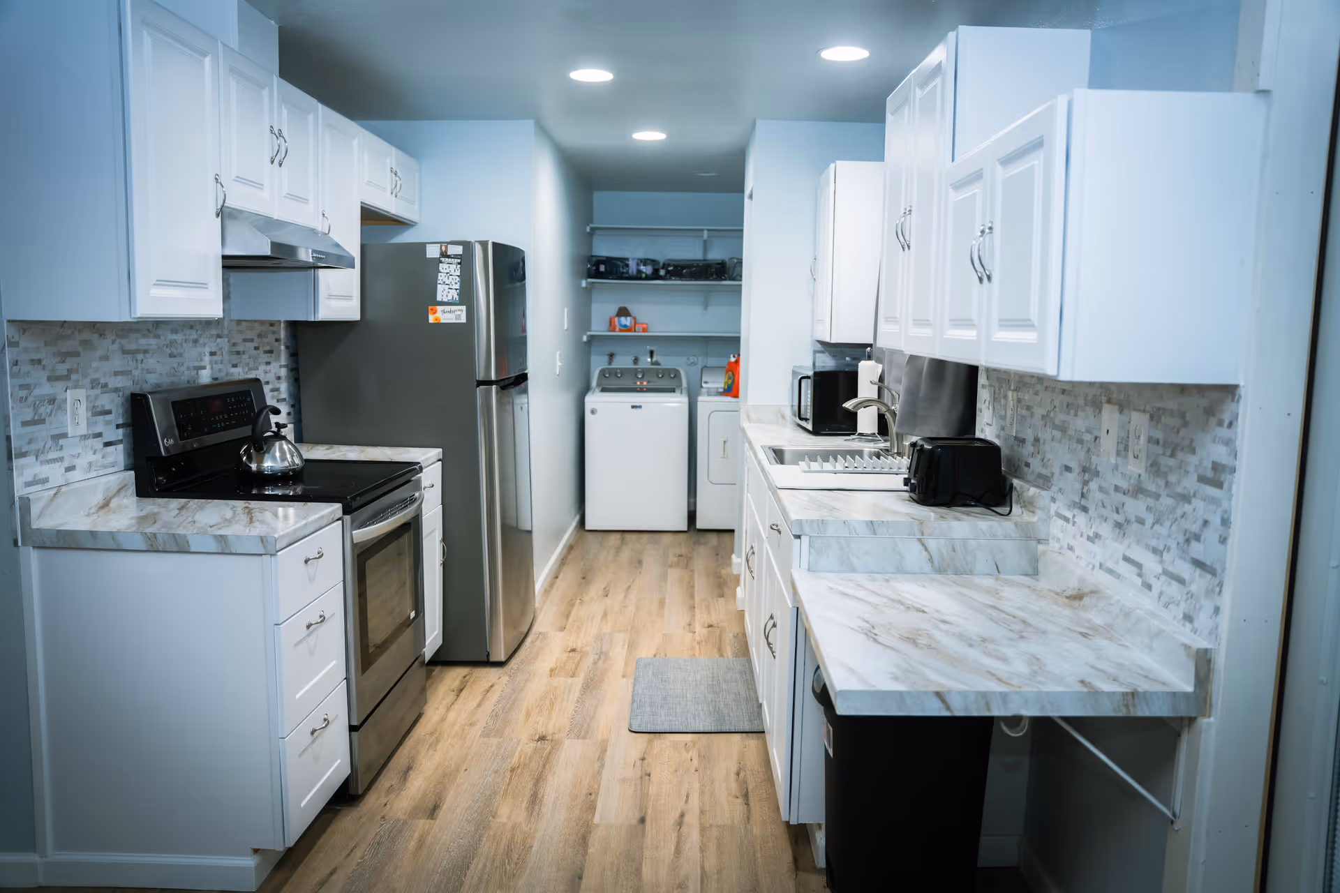 A narrow kitchen with white cabinets and marble-patterned countertops on both sides. On the left side, there is a stainless steel stove with a kettle on it and a refrigerator. On the right side, there is a sink, a microwave, a toaster, and a dish drying rack. At the far end of the kitchen, there is a laundry area with a washing machine and a dryer, along with some shelves holding laundry supplies. The floor is wooden, and the ceiling has recessed lighting.