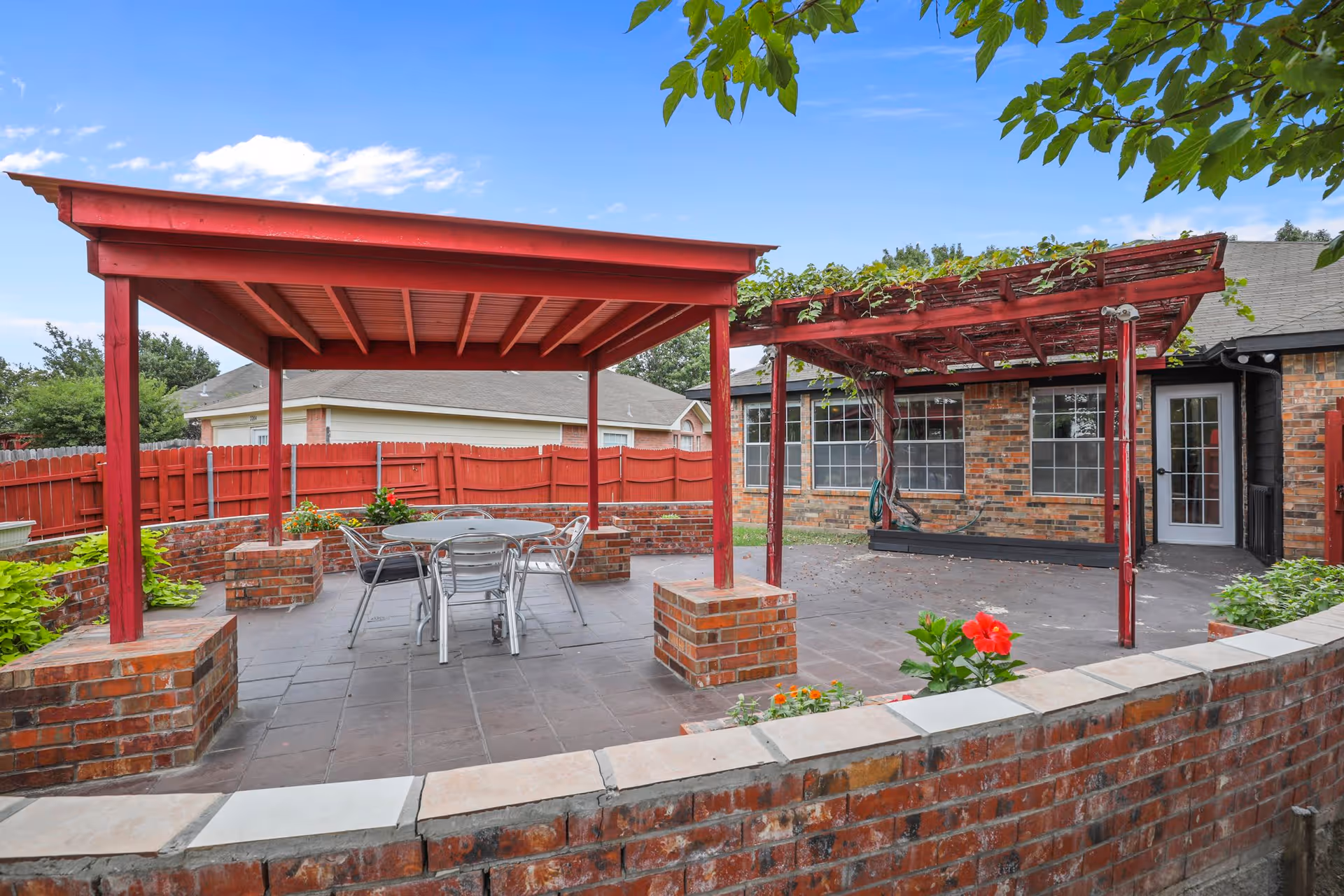Outdoor patio area with a red wooden pergola and a covered seating area. There is a round metal table with four chairs underneath the covered structure. The patio is surrounded by a low brick wall and a red wooden fence. Green plants and flowers are visible around the patio, with a brick building and windows in the background.