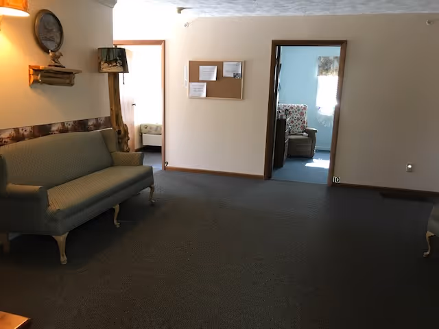Interior view of a common area in an assisted living facility with a green upholstered sofa on the left, a wall-mounted lamp above it, a clock on a small wooden shelf, and two doorways leading to rooms with chairs and windows letting in natural light.