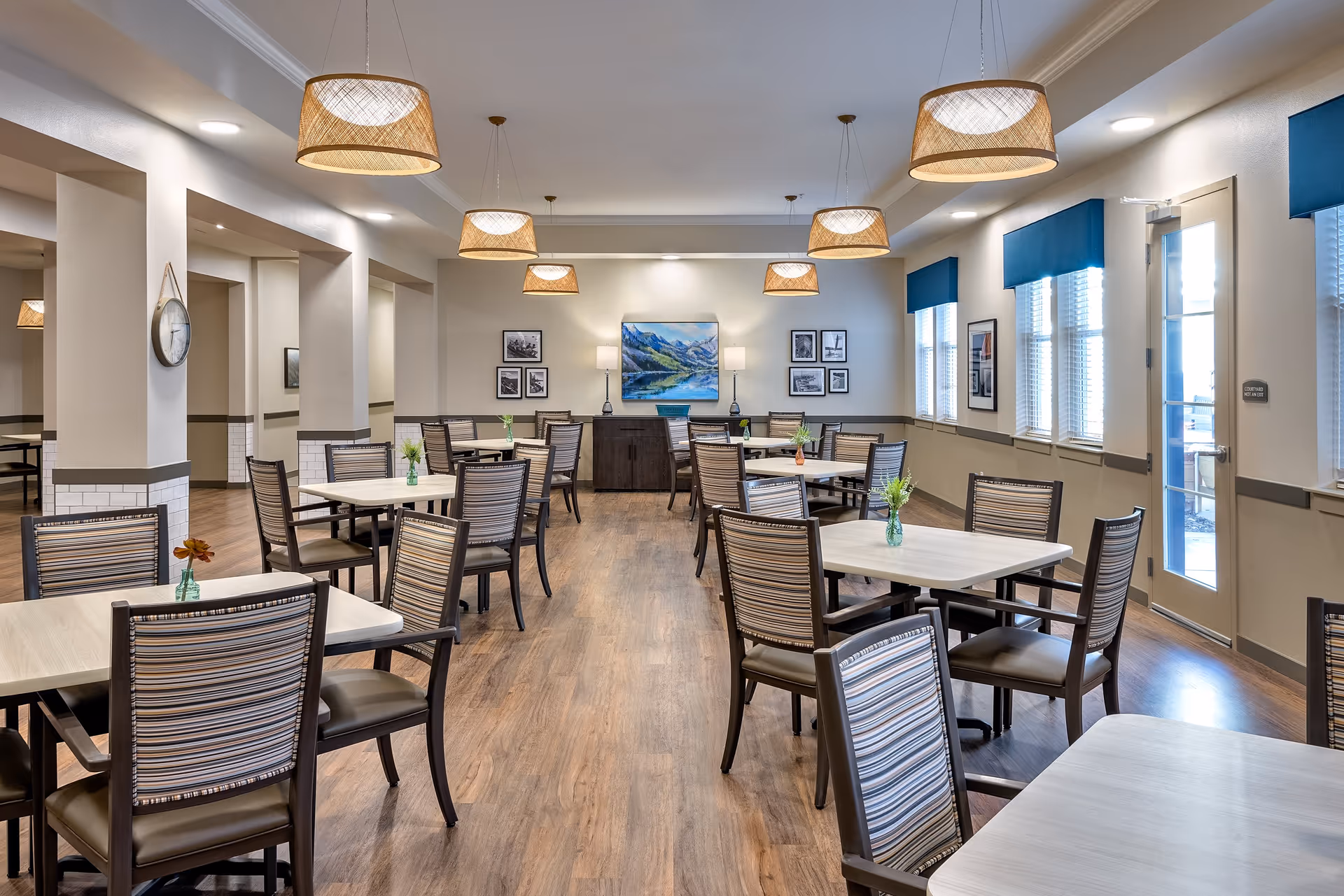 Bright communal dining room with multiple tables and chairs, pendant lights, wood flooring, and artwork on the far wall.