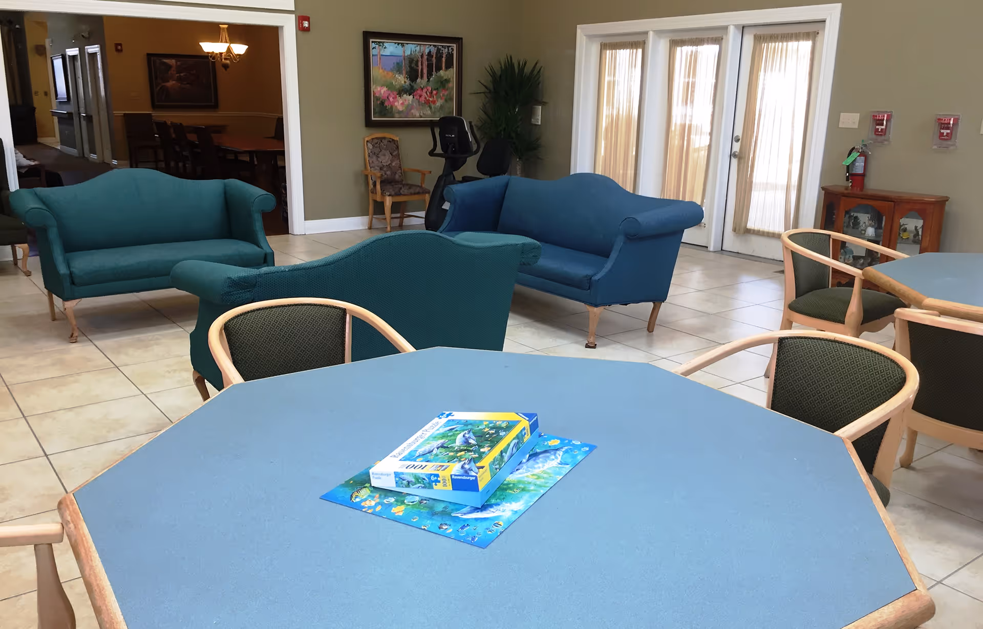 A common area in a senior living facility with blue and green upholstered sofas, a blue octagonal table with a puzzle box on it, several chairs around the table, a painting on the wall, an exercise bike, and glass doors with sheer curtains letting in natural light.