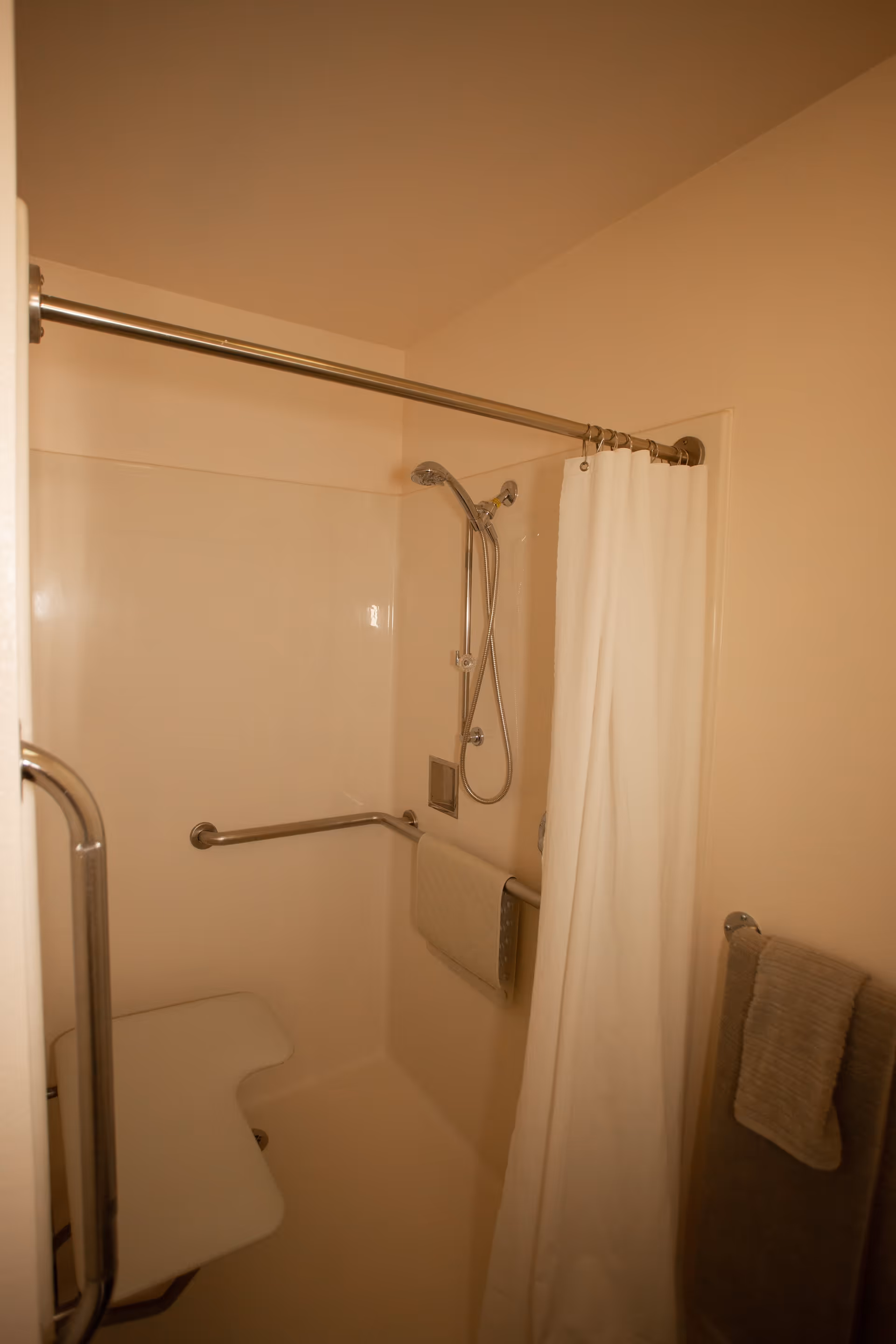 A beige-colored shower area with a white shower curtain partially drawn. The shower has a handheld showerhead attached to a metal bar, a foldable white shower seat, and metal grab bars for support. Two towels hang on the wall outside the shower.