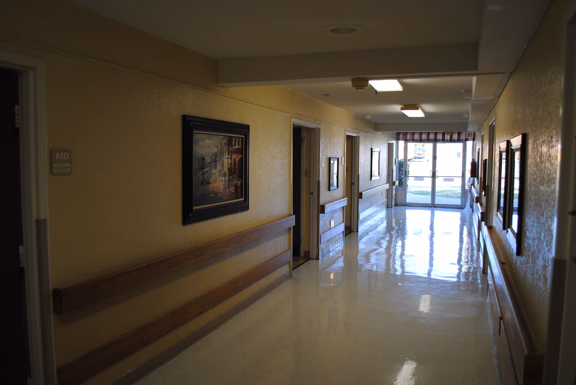 A clean, well-lit hallway in a healthcare facility with polished floors, wooden handrails on both sides, several doorways along the corridor, framed paintings on the walls, and a glass exit door at the far end letting in natural light.