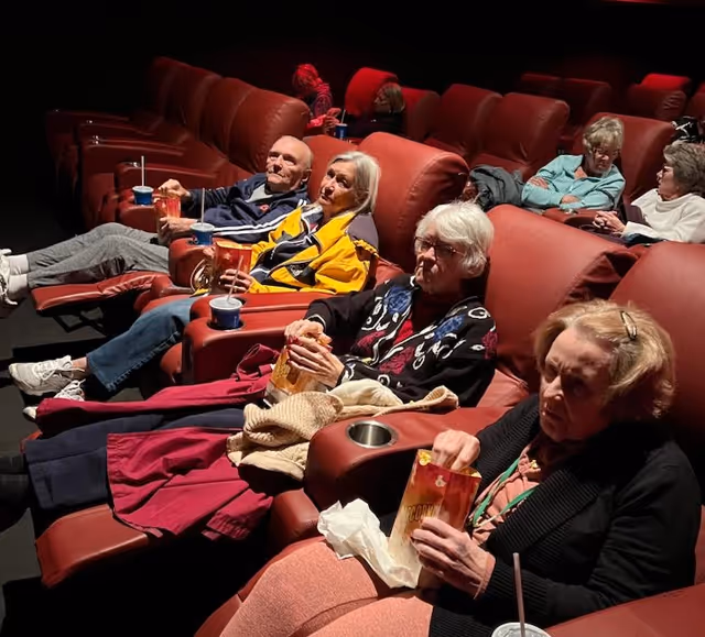 A group of elderly people sitting in red reclining theater seats, eating popcorn and drinking from cups, watching a movie in a dimly lit theater room.