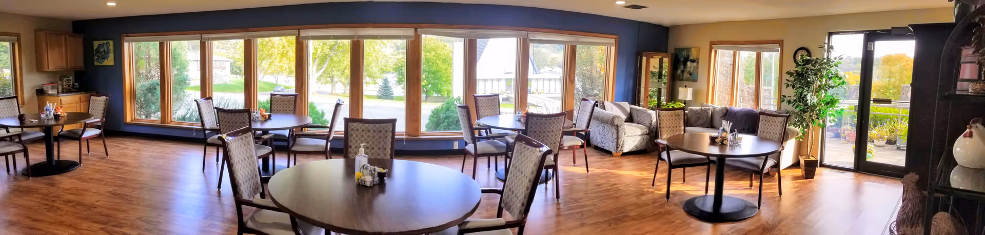 A bright dining room with multiple round tables and chairs arranged neatly. Large windows along one wall provide a view of green trees and outdoor scenery. There is a comfortable seating area with sofas near the windows and a glass door leading outside. The room has wooden flooring and is decorated with plants and artwork.
