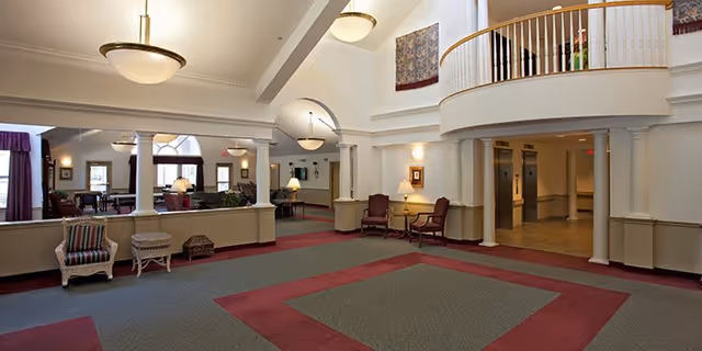 Spacious and well-lit interior common area of Randolph Village featuring high ceilings, large windows with purple curtains, multiple seating areas with chairs and lamps, decorative columns, and a balcony overlooking the space.