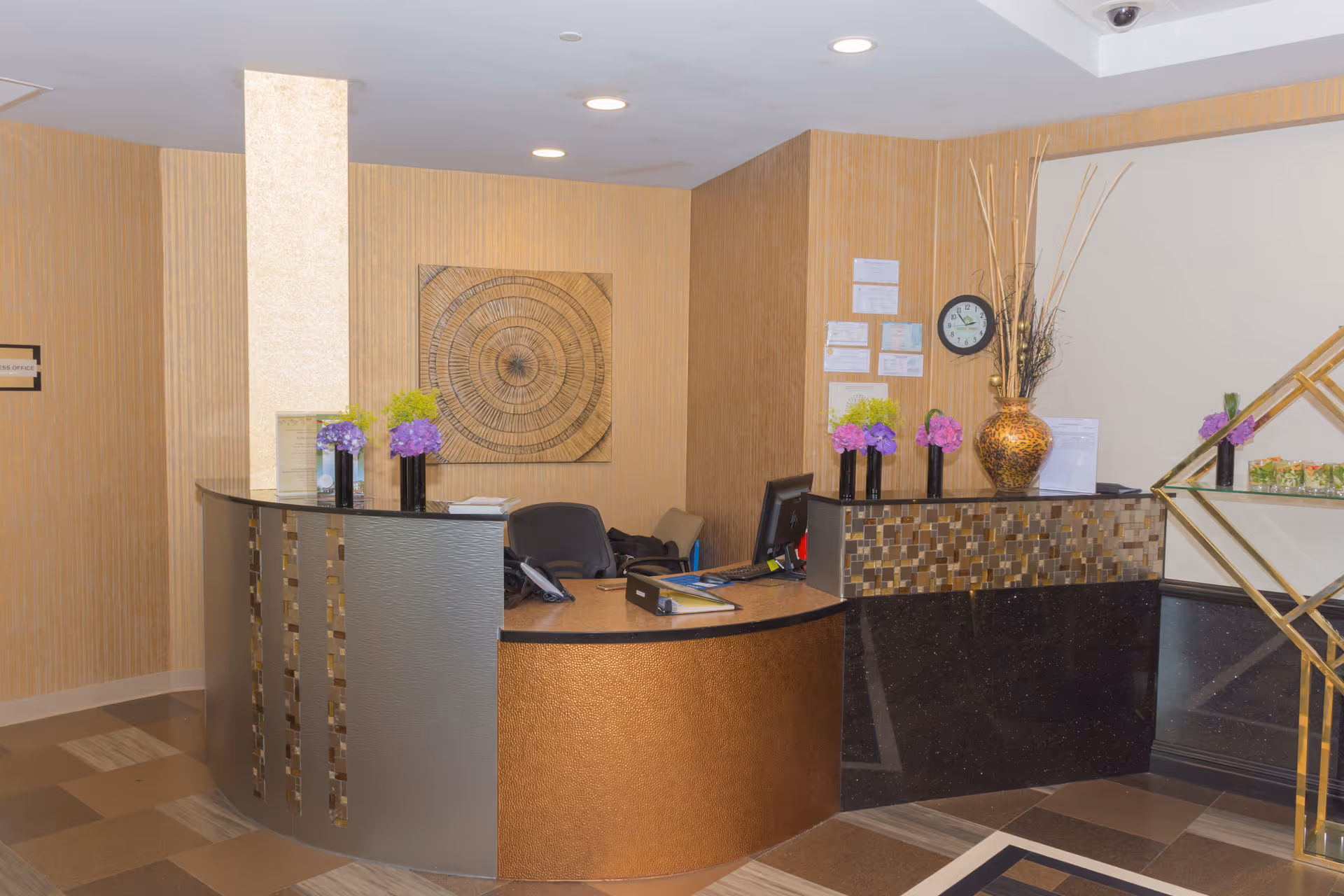 Reception desk area inside Somerset Woods Rehabilitation & Nursing Center with a curved counter, decorative vases with purple and green flowers, a wall clock, and a piece of circular wall art behind the desk.