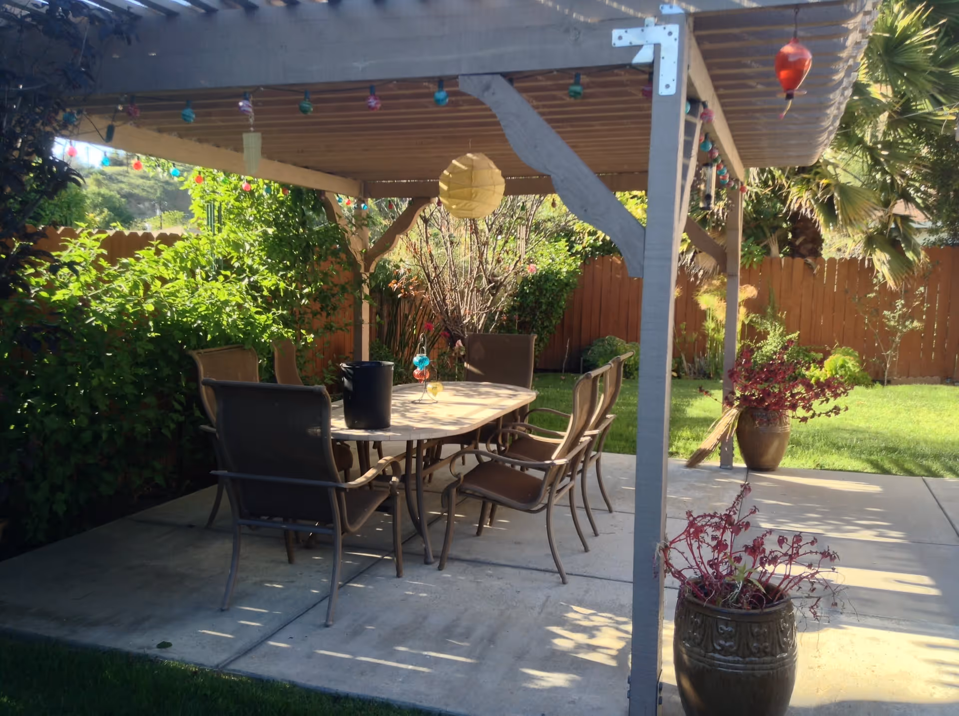 Outdoor patio area with a wooden pergola overhead, decorated with colorful string lights and a hanging paper lantern. Underneath is a rectangular table surrounded by six chairs. The patio is surrounded by green bushes, trees, and a wooden fence. There are two large decorative pots with plants on the concrete floor.