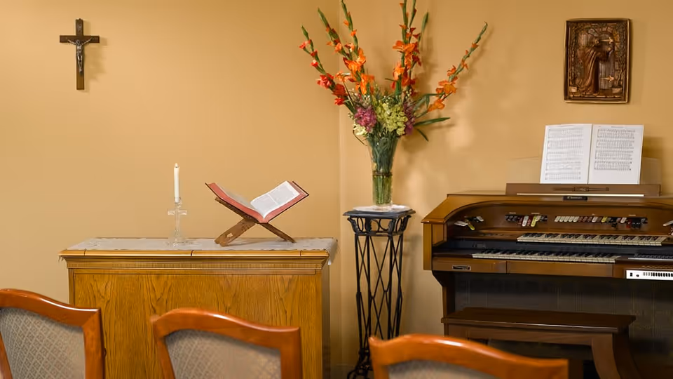 A small chapel or prayer room with a wooden cabinet holding an open book on a stand and a lit candle in a glass holder. A crucifix hangs on the wall above the cabinet. To the right, there is a tall vase with colorful flowers on a decorative metal stand and an organ with sheet music on the stand. Several wooden chairs with cushioned backs are partially visible in the foreground.