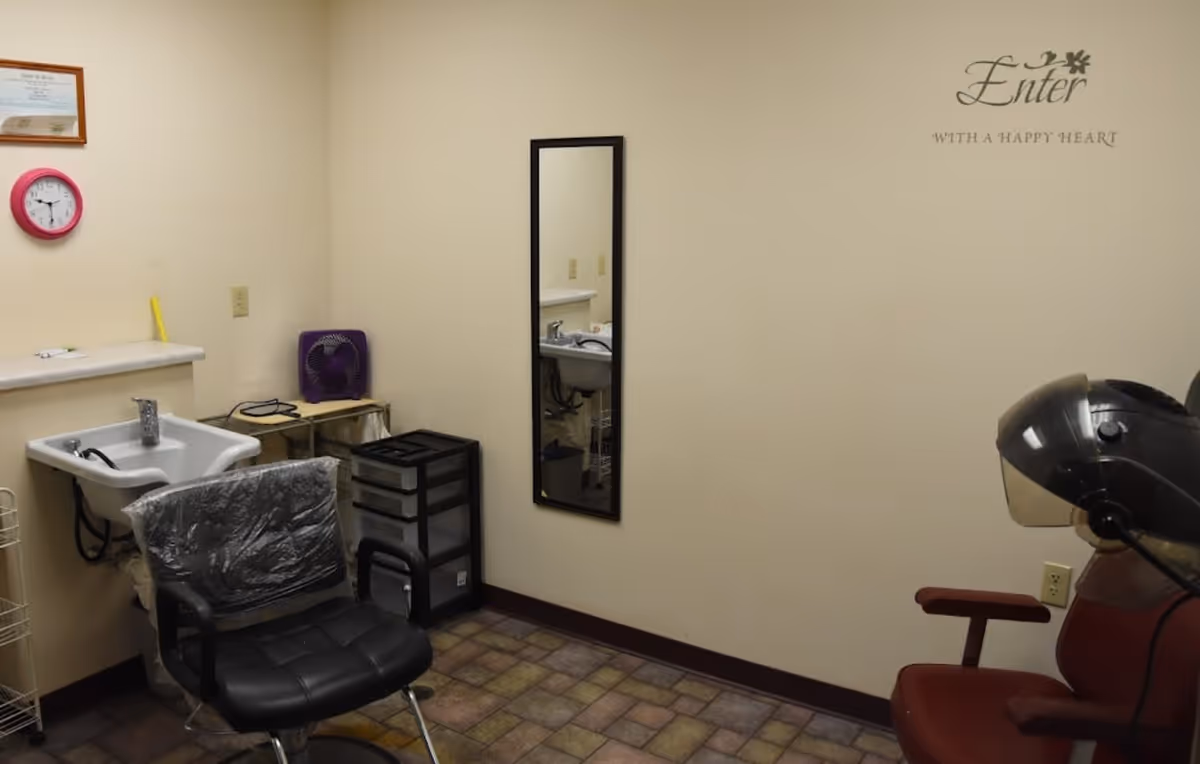 Interior of a small salon or grooming room with a black salon chair covered in plastic, a white sink, a purple fan on a small table, a black storage drawer unit, a wall-mounted mirror, and a red salon chair with a hair dryer hood. The walls are beige with a decorative wall decal that reads 'Enter with a happy heart'.