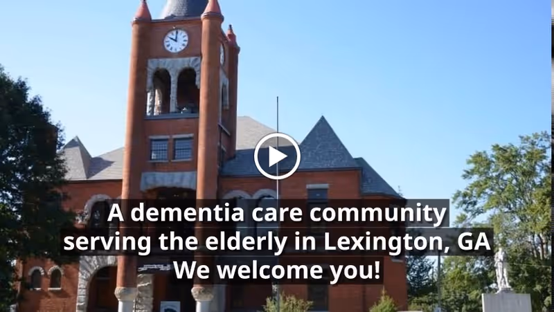 Exterior view of a large brick building with a clock tower under a clear blue sky, surrounded by trees. Text overlay reads: 'A dementia care community serving the elderly in Lexington, GA We welcome you!'