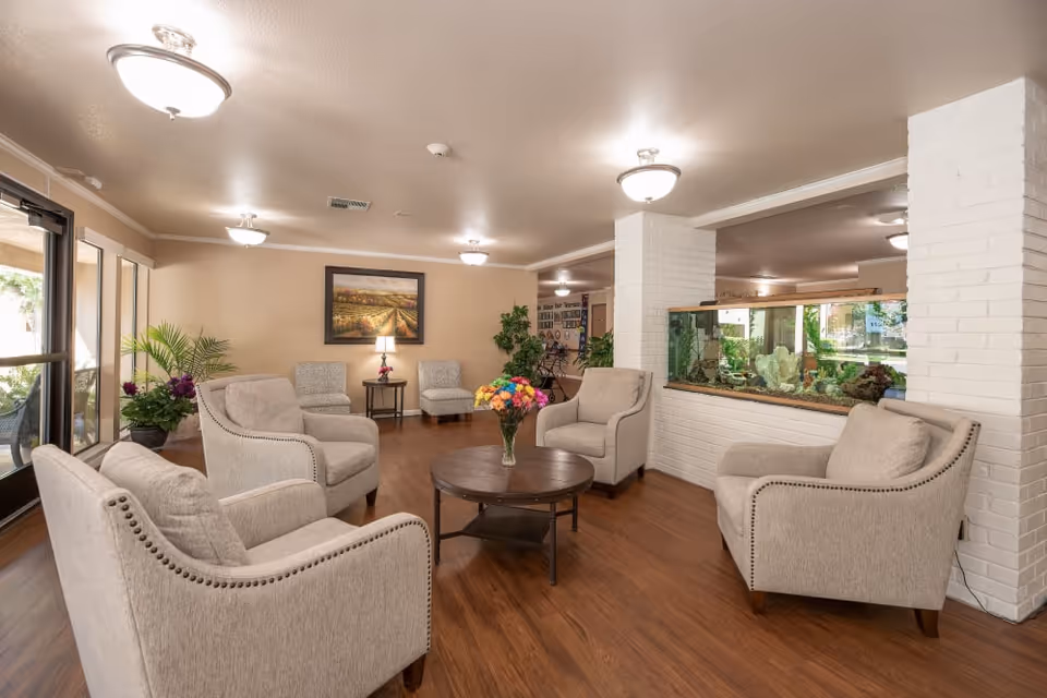 A cozy senior living facility common area with six beige armchairs arranged around a round wooden coffee table with a vase of colorful flowers. The room features wood flooring, white brick pillars, a large aquarium built into one pillar, and a framed landscape painting on the wall. There are ceiling lights and large windows letting in natural light.