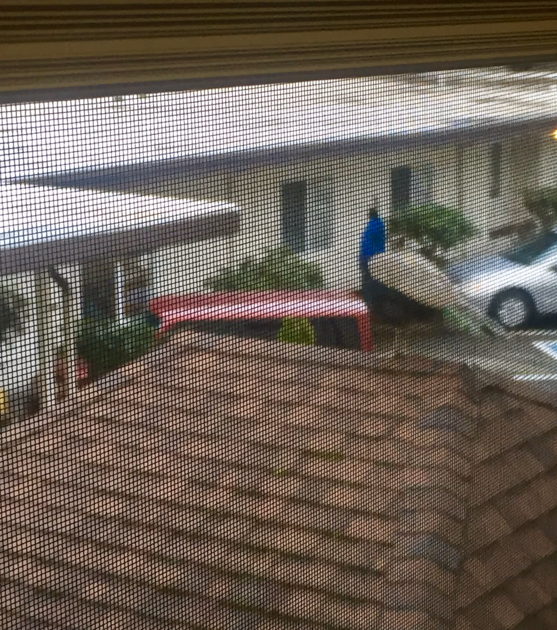 View through a window screen of an exterior courtyard showing building roofs, a parked car, a red bench, and a person in a blue jacket near shrubbery.