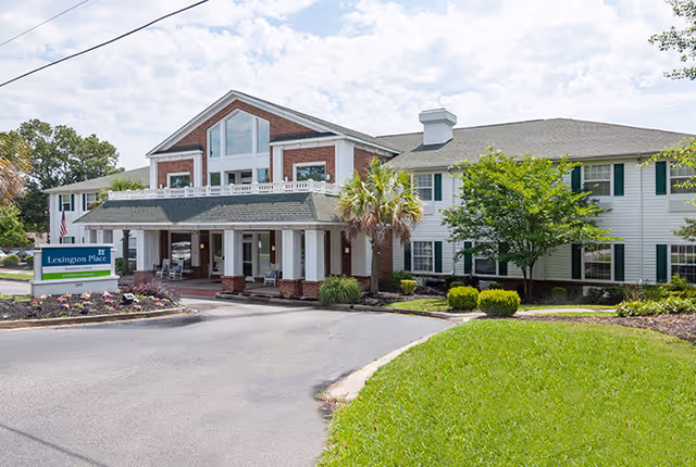 Exterior view of a two-story assisted living facility building with a covered entrance, surrounded by greenery and a well-maintained lawn under a partly cloudy sky.