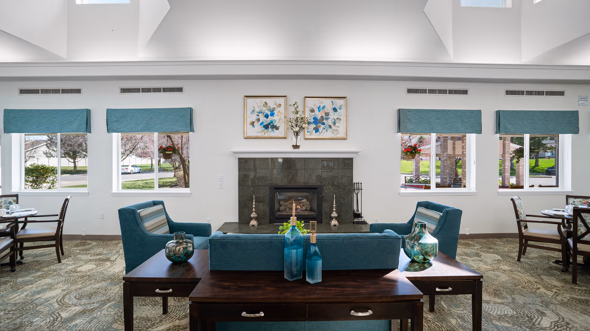 A bright and spacious common area in a senior living facility featuring a seating arrangement with teal upholstered chairs and a matching sofa around a dark wooden coffee table. The room has large windows with teal valances letting in natural light, a fireplace with decorative items on the mantel, and framed floral artwork above it. There are dining tables and chairs visible on both sides of the seating area, and the carpet has a patterned design.