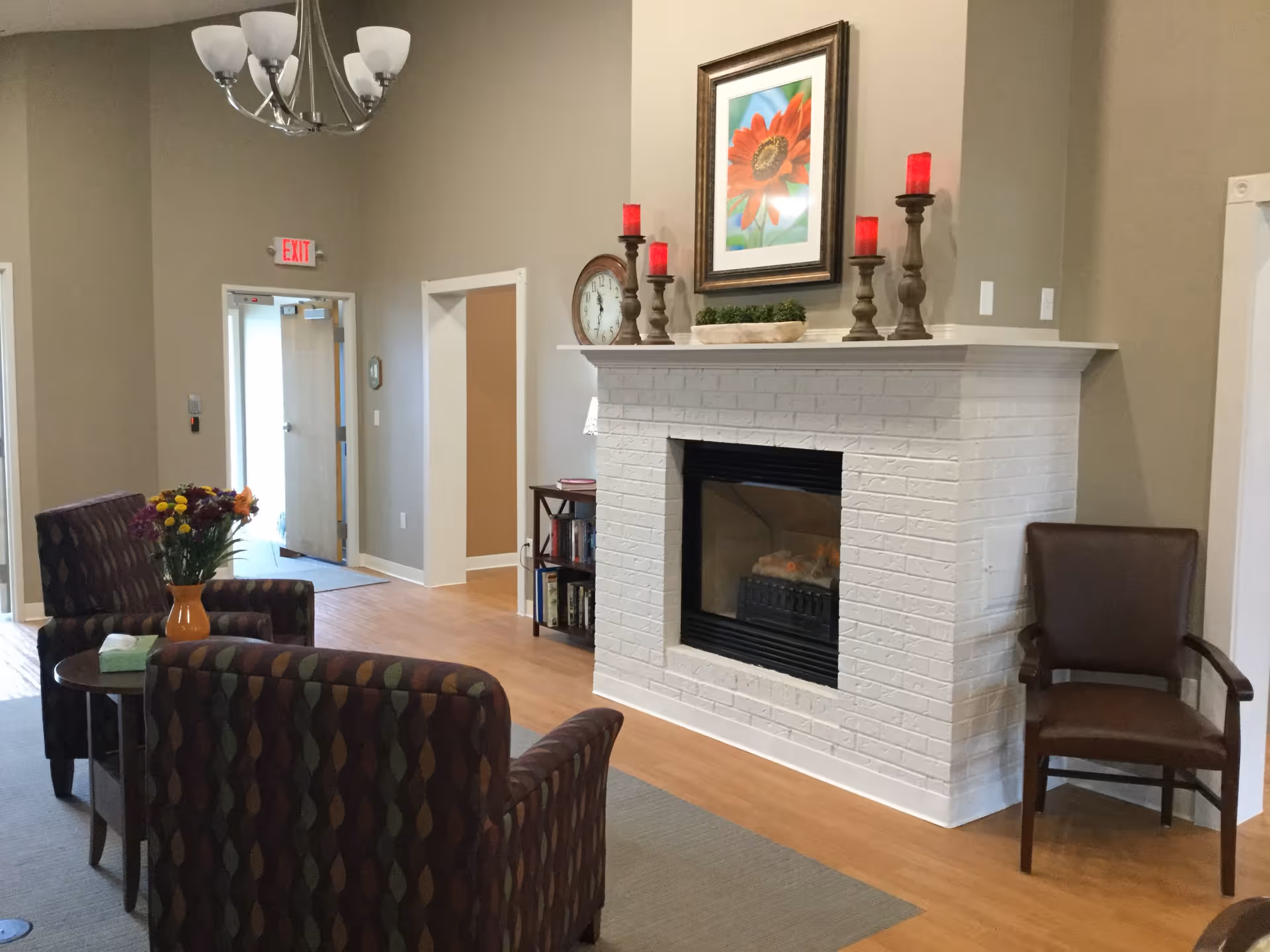A cozy living area in a senior living facility featuring a white brick fireplace with red candles and a framed flower picture above it. Two patterned armchairs face a small round table with a vase of flowers and a box of tissues. A brown wooden chair is placed near the fireplace. The room has light wood flooring, beige walls, and a chandelier hanging from the ceiling. An exit door is visible in the background.