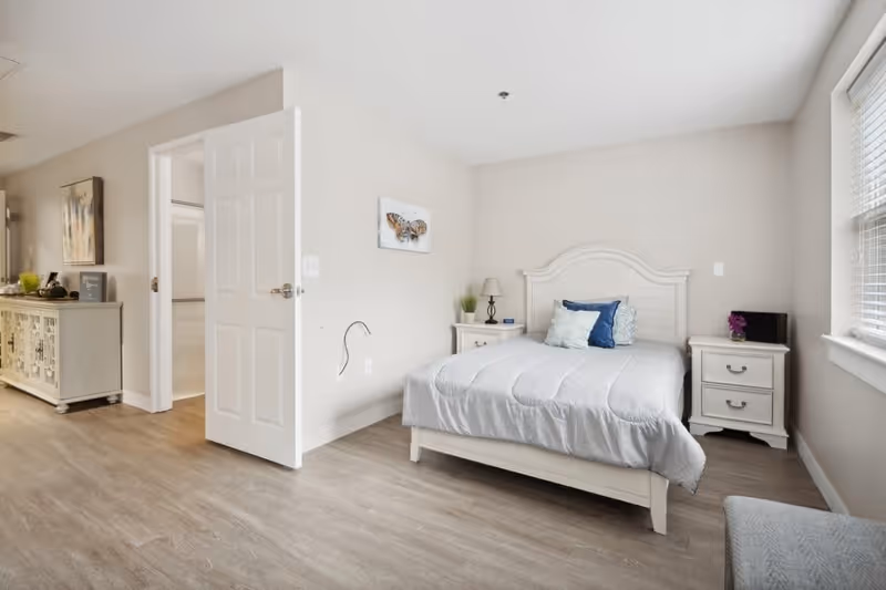 A bright bedroom with a white wooden bed frame and a neatly made bed with white and blue pillows. There are two white nightstands on either side of the bed, one with a lamp and a small plant, and the other with a small purple flower and a black object. A window with blinds is on the right wall, allowing natural light into the room. The floor is light wood, and there is a white door open to an adjacent room with a dresser and artwork visible.