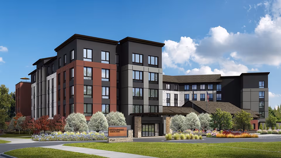 Exterior view of a modern multi-story senior living facility with a mix of dark and red paneling, large windows, landscaped greenery, and a clear blue sky with some clouds.