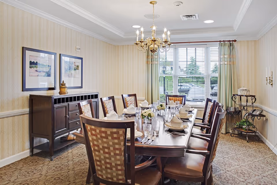 A formal dining room with a large wooden dining table set for eight people. The table is arranged with plates, bowls, glasses, napkins, and silverware. The room features patterned carpet, wallpaper with subtle vertical stripes, a chandelier hanging from the ceiling, two framed pictures on the wall, a sideboard, and a window with blinds and curtains letting in natural light.