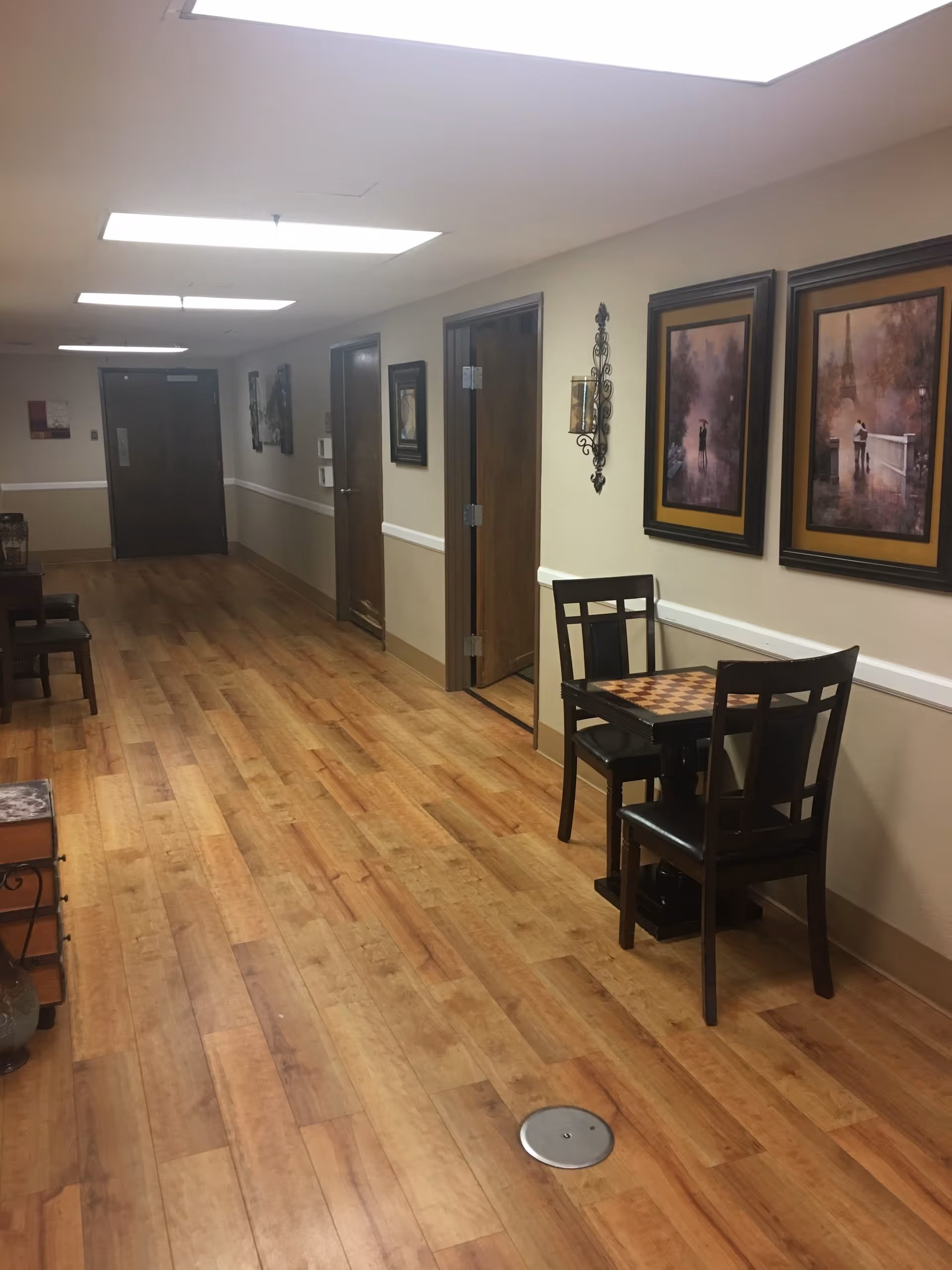 A hallway in an assisted living facility with wooden flooring and beige walls. There are several closed wooden doors along the hallway, framed artwork on the walls, and a small table with two chairs on the right side. The ceiling has rectangular fluorescent lights.