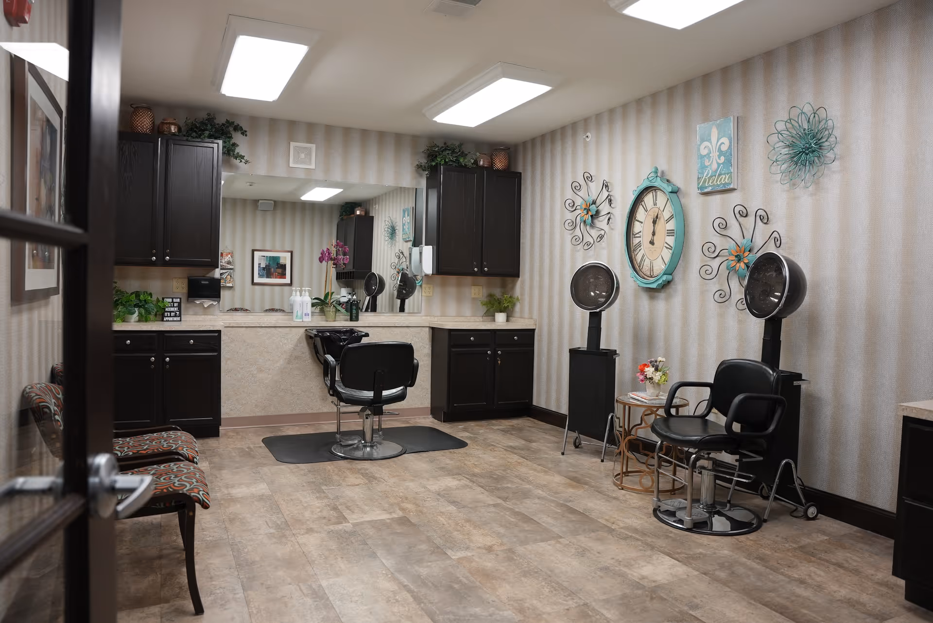 Interior salon room with styling chairs, hooded hair dryers, cabinets, and decorative wall clock and artwork.