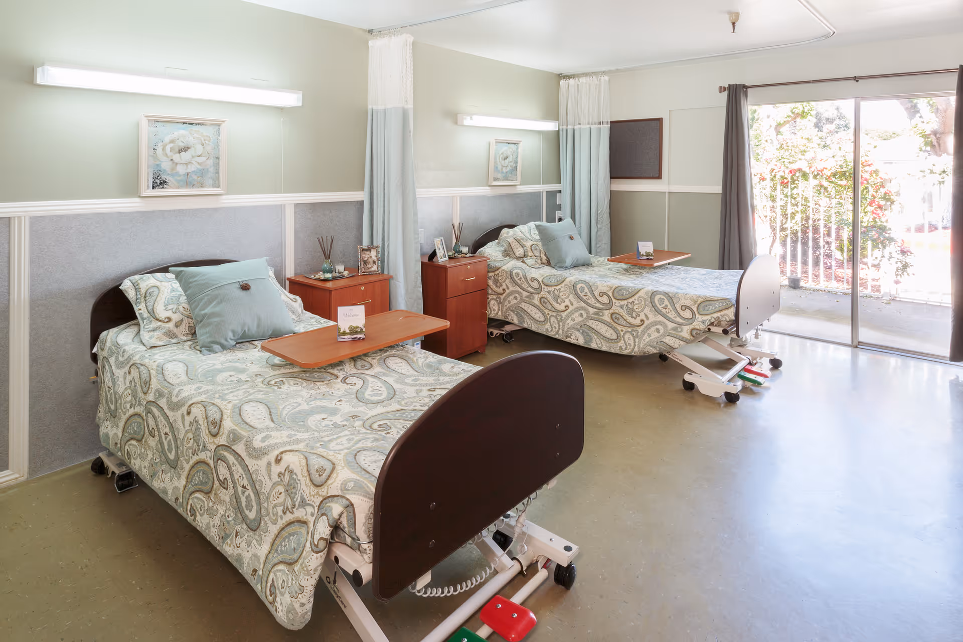 A bright and clean room with two hospital-style beds, each with patterned bedding and pillows. Each bed has a wooden overbed table and is positioned next to a small wooden nightstand with decorative items. The room has light green walls with white trim, a large window with sliding glass doors letting in natural light, and curtains that can be drawn for privacy.