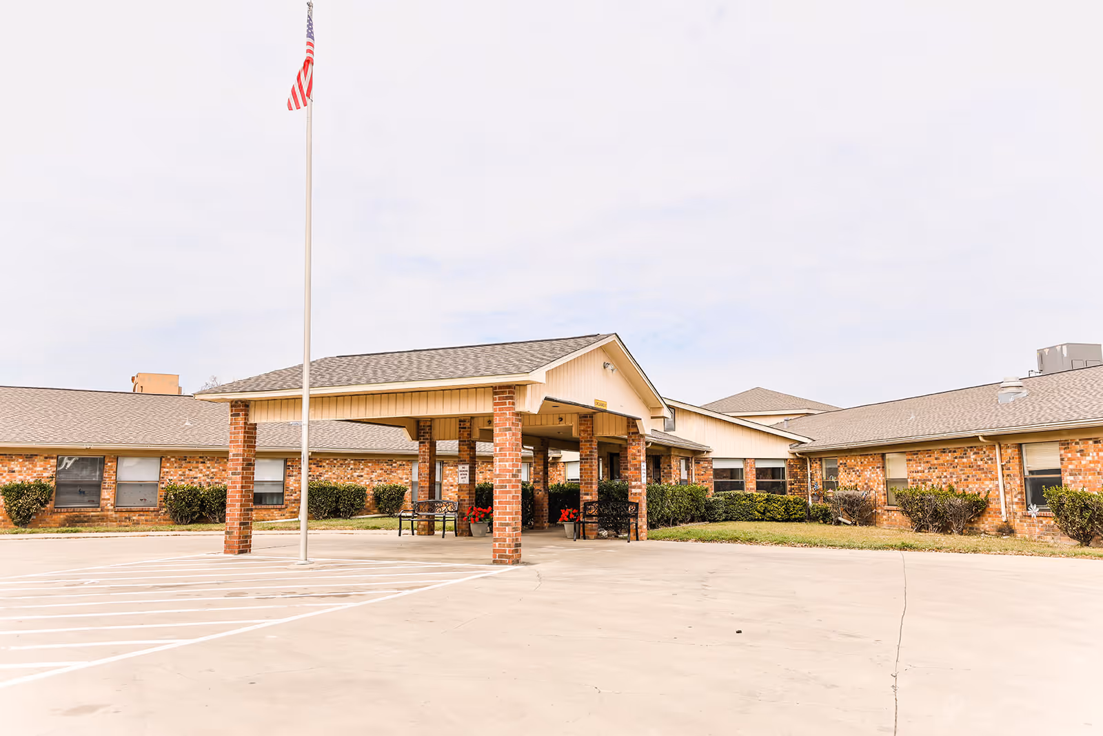 Exterior view of Lily Springs Rehabilitation and Healthcare Center showing a single-story brick building with a covered entrance supported by brick columns. There is an American flag on a flagpole in front of the entrance and some bushes along the building. The sky is overcast.