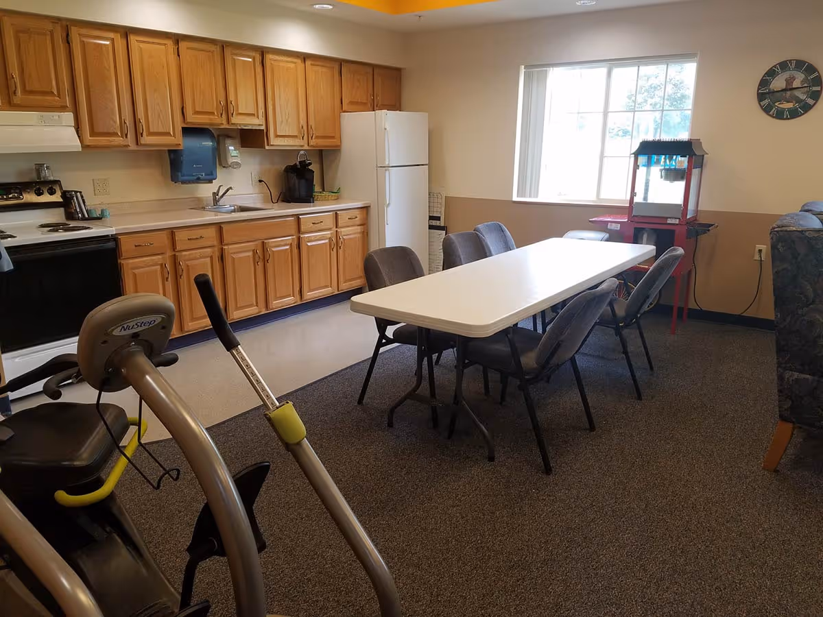 Communal dining and kitchenette area with a long folding table and chairs, wooden cabinets and appliances, and exercise equipment in the foreground.