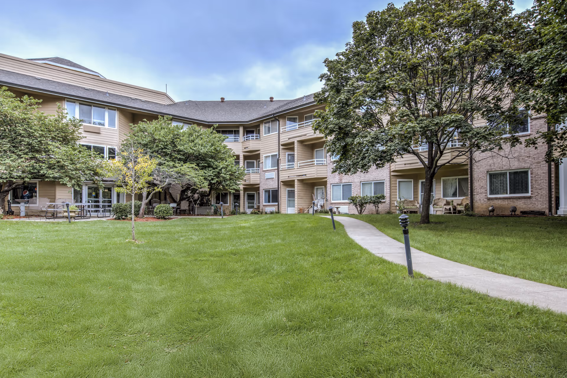 Outdoor view of a senior living facility showing a green lawn with a concrete pathway leading to a multi-story building with balconies and windows. Trees and bushes are planted around the lawn, and there are benches and outdoor seating areas near the building.