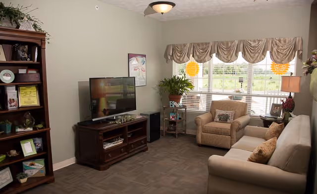 A cozy living room with a beige sofa and armchair, a wooden TV stand with a flat-screen television, a wooden bookshelf filled with books and decorative items, a small side table with a lamp, and a large window with beige valance curtains letting in natural light. There are plants and framed pictures adding a homey touch.