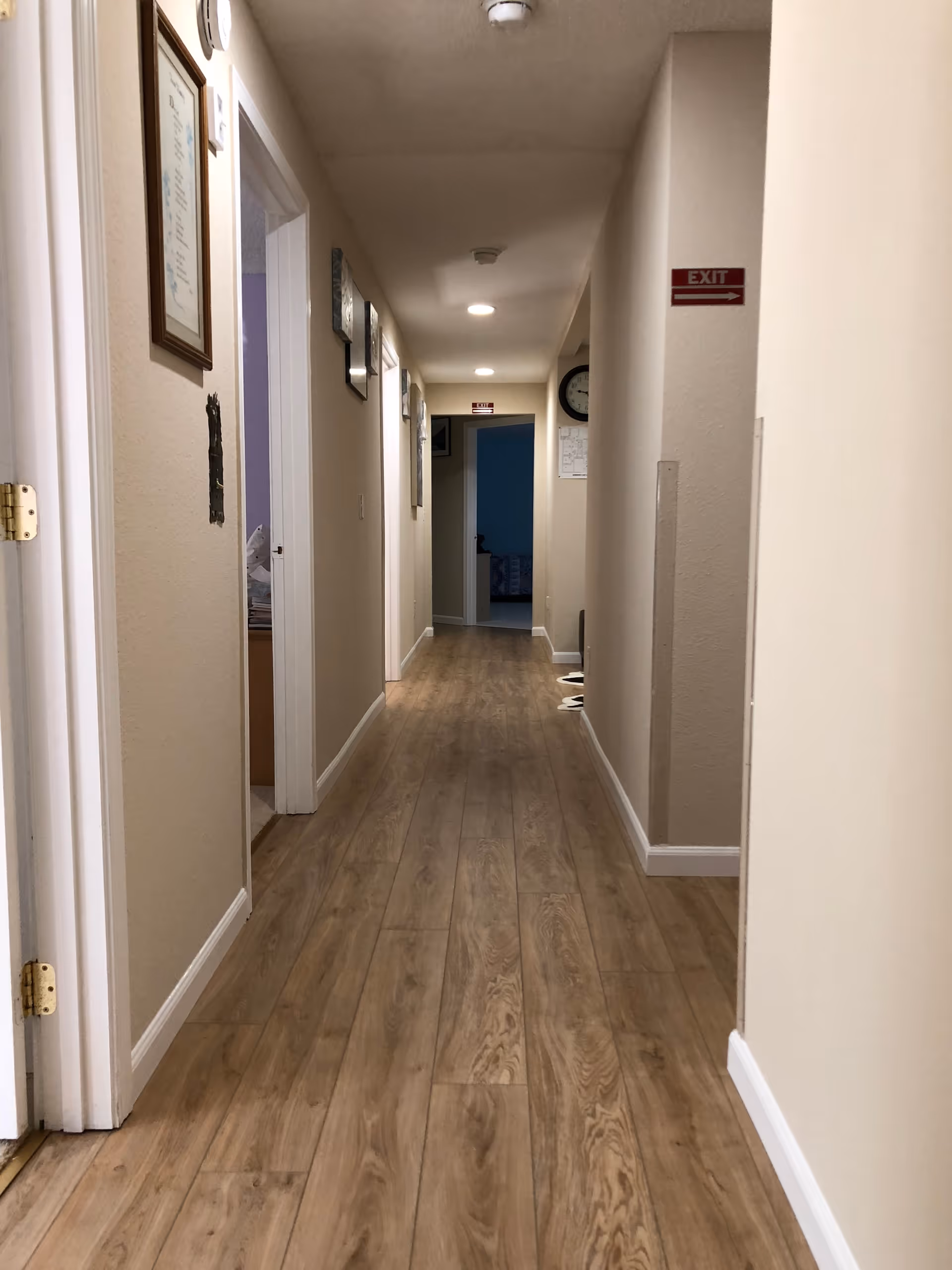A long indoor hallway with light beige walls and wood-patterned flooring. Several doorways line the left side of the hallway, and there are framed pictures and a clock on the walls. Ceiling lights illuminate the corridor, and an exit sign is visible on the right wall.
