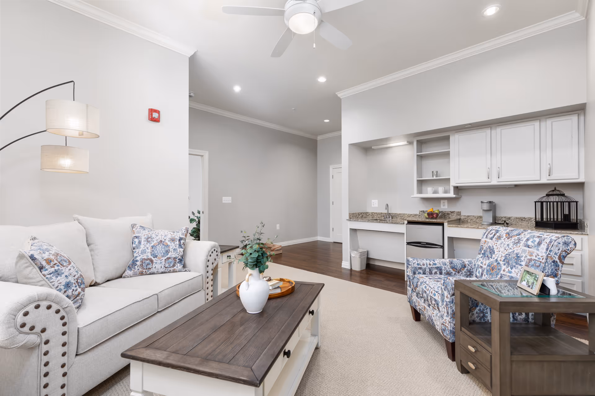 A bright and clean living room area in an assisted living facility featuring a light gray sofa with patterned cushions, a wooden coffee table with a white vase and tray, a patterned armchair next to a wooden side table with a framed photo, a ceiling fan, and a kitchenette with granite countertops, white cabinets, a small refrigerator, and a coffee maker.