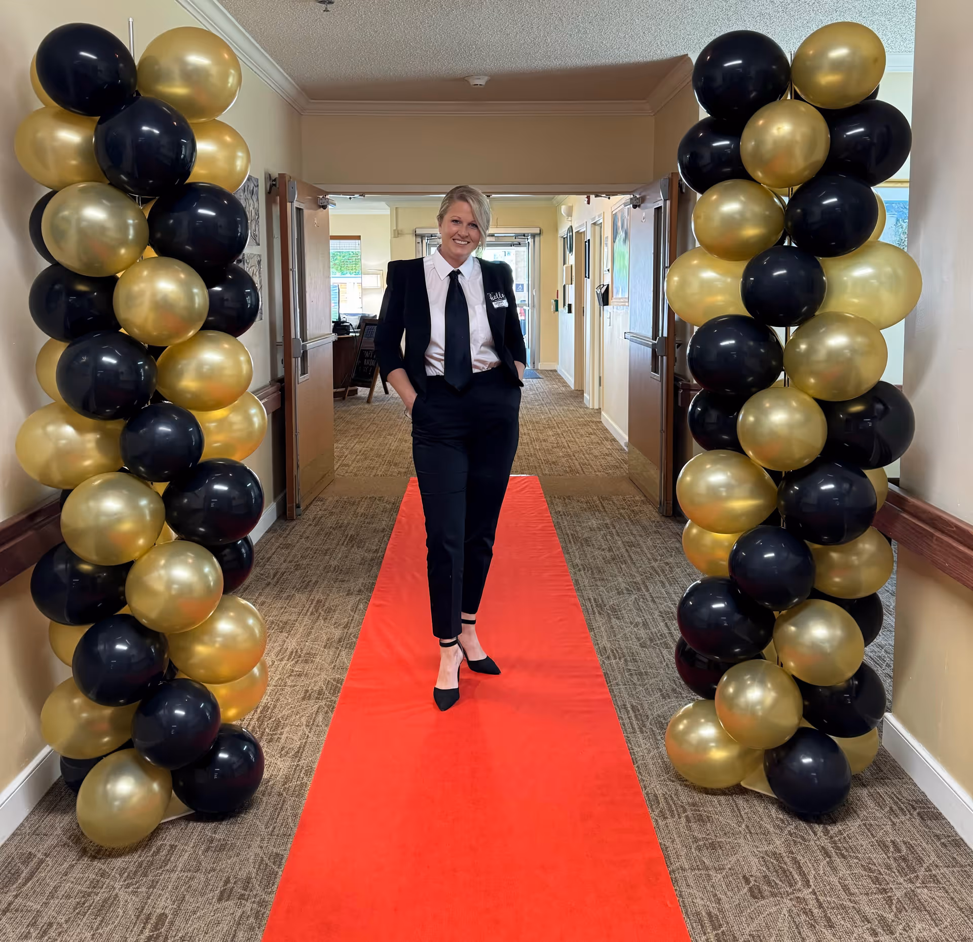 A smiling staff member stands on a red carpet in a hallway flanked by black and gold balloon columns.